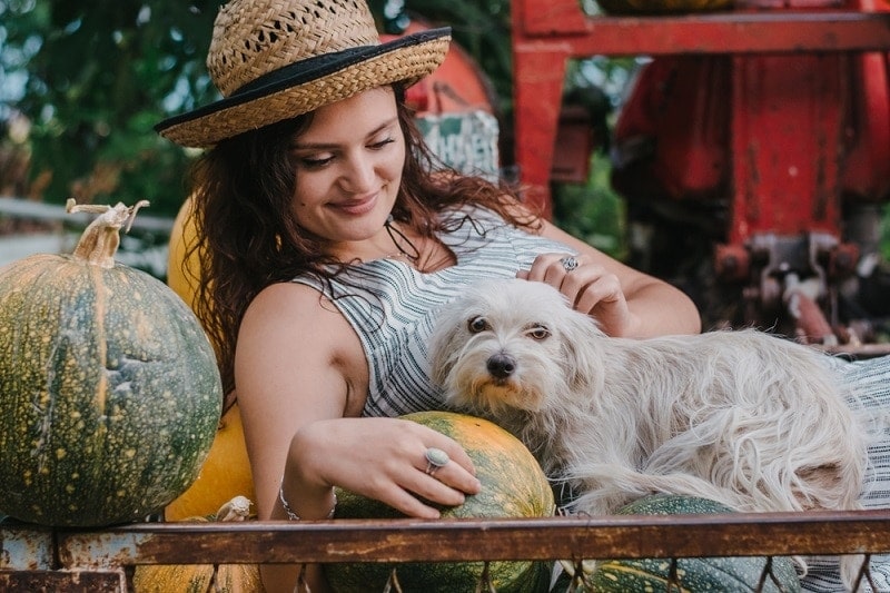 Young countryside woman cuddling her dog while lying among pumpkins