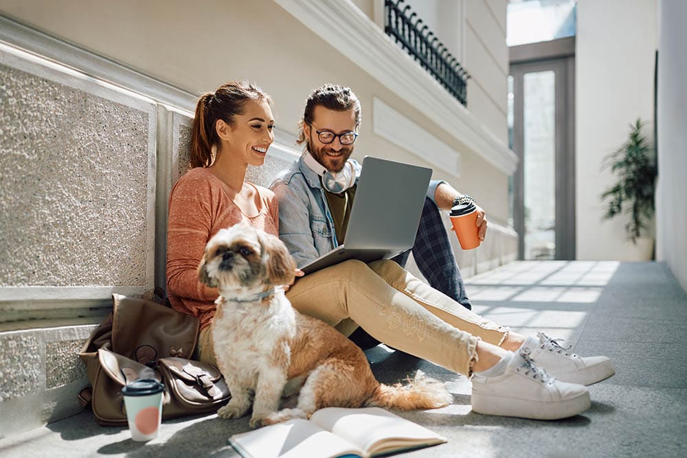 Young happy students e-learning over laptop while sitting in a hallway with their dog insurance