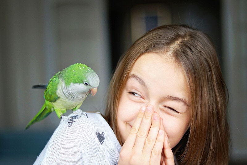 Cute smiling girl playing with her pet green Monk Parakeet parrot