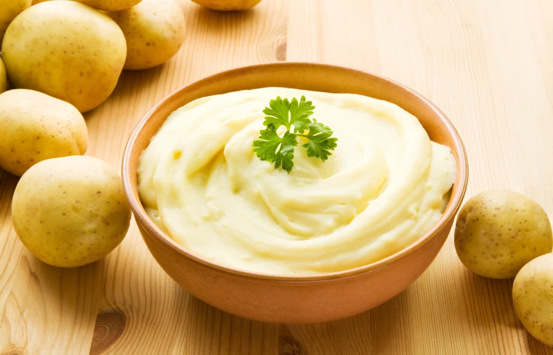 bowl of mashed potatoes surrounded with uncooked potatoes on wooden surface