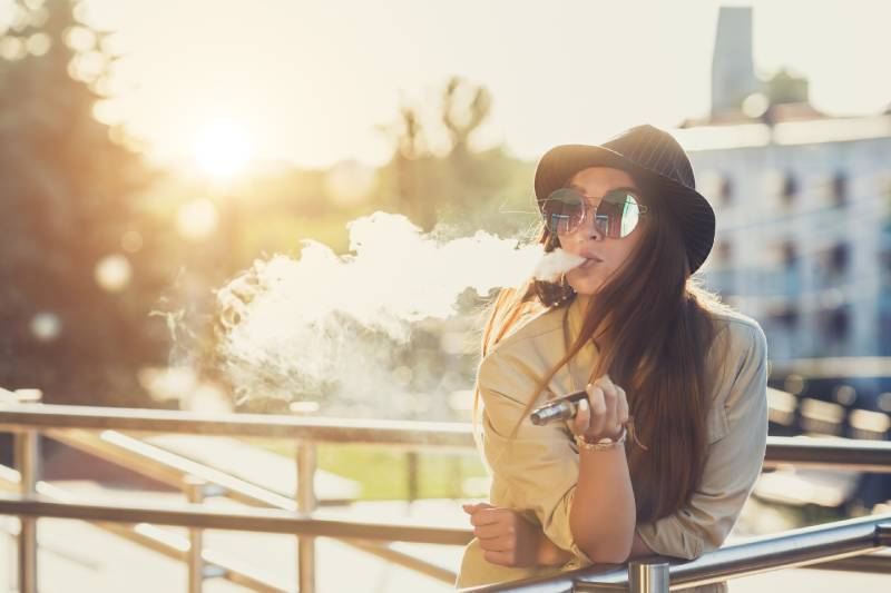 Pretty young hipster woman in black hat vape ecig, vaping device at the sunse